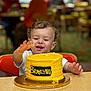 Braylynn joined the competition — help win amazing prizes! toddler, child, cake, yellow_cake, birthday_cake, table, high_chair, smiling, happy, food, icing, celebration, indoor, person, curly_hair, frosting, hand, face, party, dessert