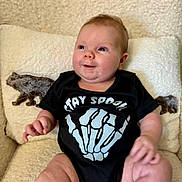 Riley joined the competition — help win amazing prizes! baby, infant, child, onesie, black_clothing, skeleton_hand, text_print, chair, cushion, bear_design, cozy, comfortable, smiling, happy, cute, indoors, portrait, soft_texture, chubby, sitting