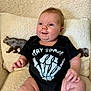 baby, infant, child, onesie, black_clothing, skeleton_hand, text_print, chair, cushion, bear_design, cozy, comfortable, smiling, happy, cute, indoors, portrait, soft_texture, chubby, sitting