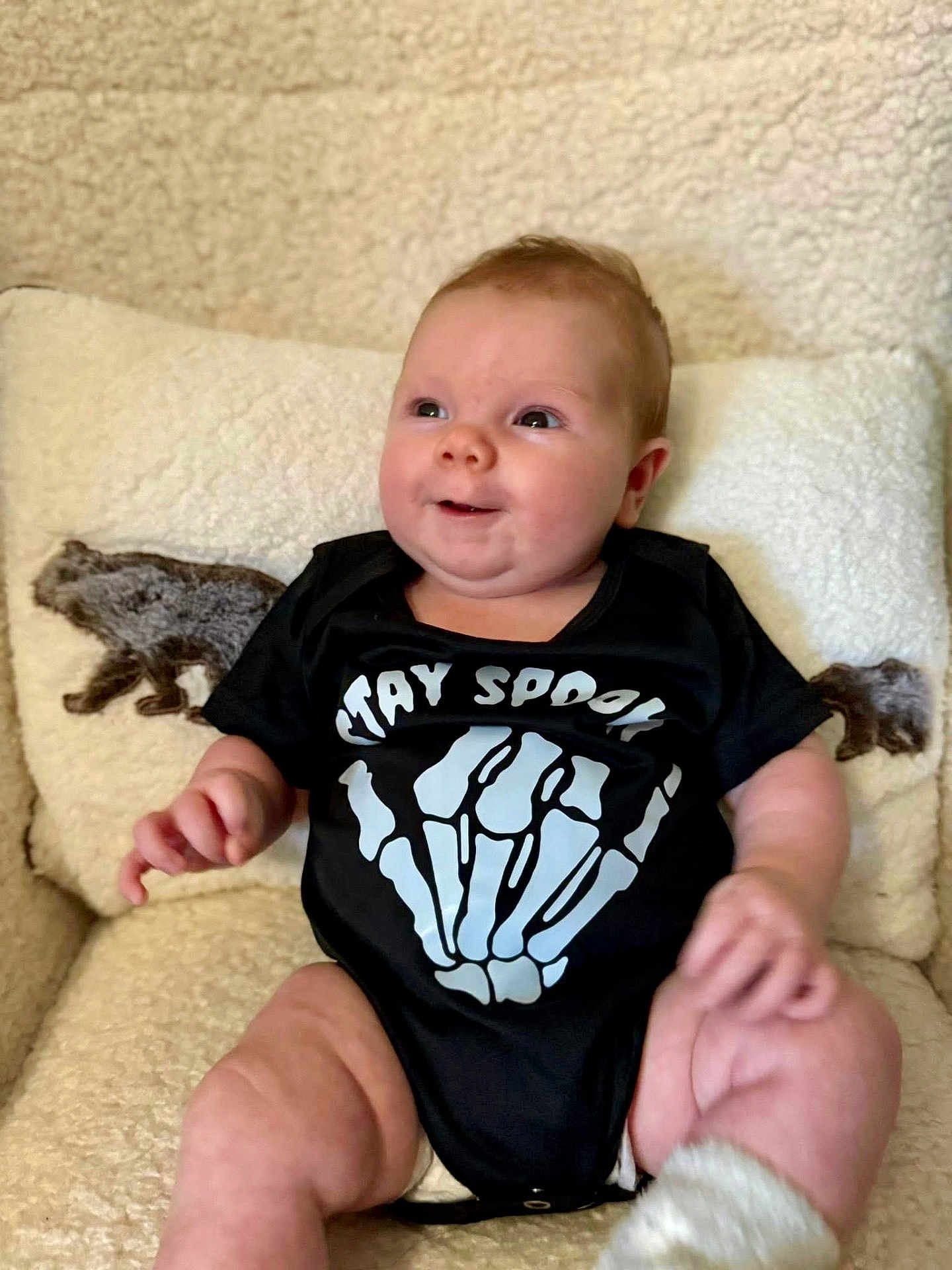 Riley joined the competition — help win amazing prizes! baby, infant, child, onesie, black_clothing, skeleton_hand, text_print, chair, cushion, bear_design, cozy, comfortable, smiling, happy, cute, indoors, portrait, soft_texture, chubby, sitting
