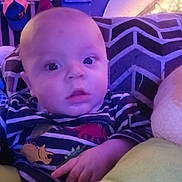 Jordan is registered to the contest to win money with this photo: baby, infant, portrait, close_up, indoor, striped_clothing, dinosaur_print, couch, cushion, pillow, surprised_expression, big_eyes, hand, bald_head, face, soft_lighting, purple_tint, blanket, home, cozy