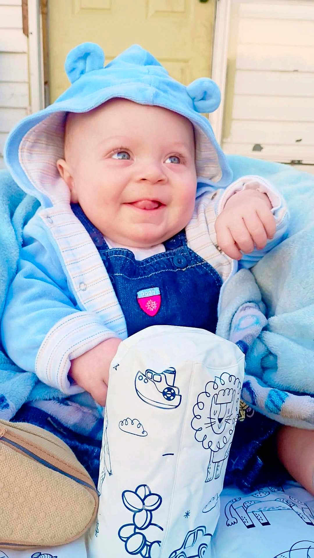 Jordan is registered to the contest to win money with this photo: baby, infant, smiling_baby, blue_hood, hood, overalls, blanket, toy, sitting, portrait, outdoor, door, cute, happy, tongue_out, eyes, hand, plush, fabric, seat