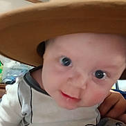 Jordan is registered to the contest to win money with this photo: adult_hand, baby, bedroom, big_eyes, blurred_background, brim, candid, ceiling, close_up, clothing, face, hand, hat, indoor, infant, onesie, portrait, rosy_cheeks, smile, storage_box