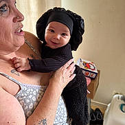 Arya a rejoint le concours — aidez-le/la à gagner de superbes lots ! arm, baby, black_outfit, bonnet, box, clothing, face, floor, furniture, happy, holding, home, indoor, light, patterned_clothing, person, shoes, smile, tattoo, woman