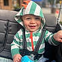 baby, blue_eyes, blurred_background, child, clothing, cute, face, hand, happy, hoodie, orange_spikes, outdoor, person, portrait, seatbelt, sippy_cup, smiling, striped_clothing, stroller, toddler
