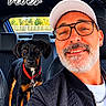 man, person, dog, black_dog, tan_markings, car, car_interior, selfie, portrait, glasses, hat, beard, smiling, red_collar, leash, seated, outdoor, sunlight, good_vibes, music_overlay