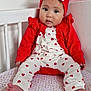 baby, barefoot, bow, child, crib, curious_expression, cute, floral_sheet, headband, heart_pattern, indoors, infant, nursery, person, portrait, red_clothing, sitting, soft_lighting, white_clothing, young_child