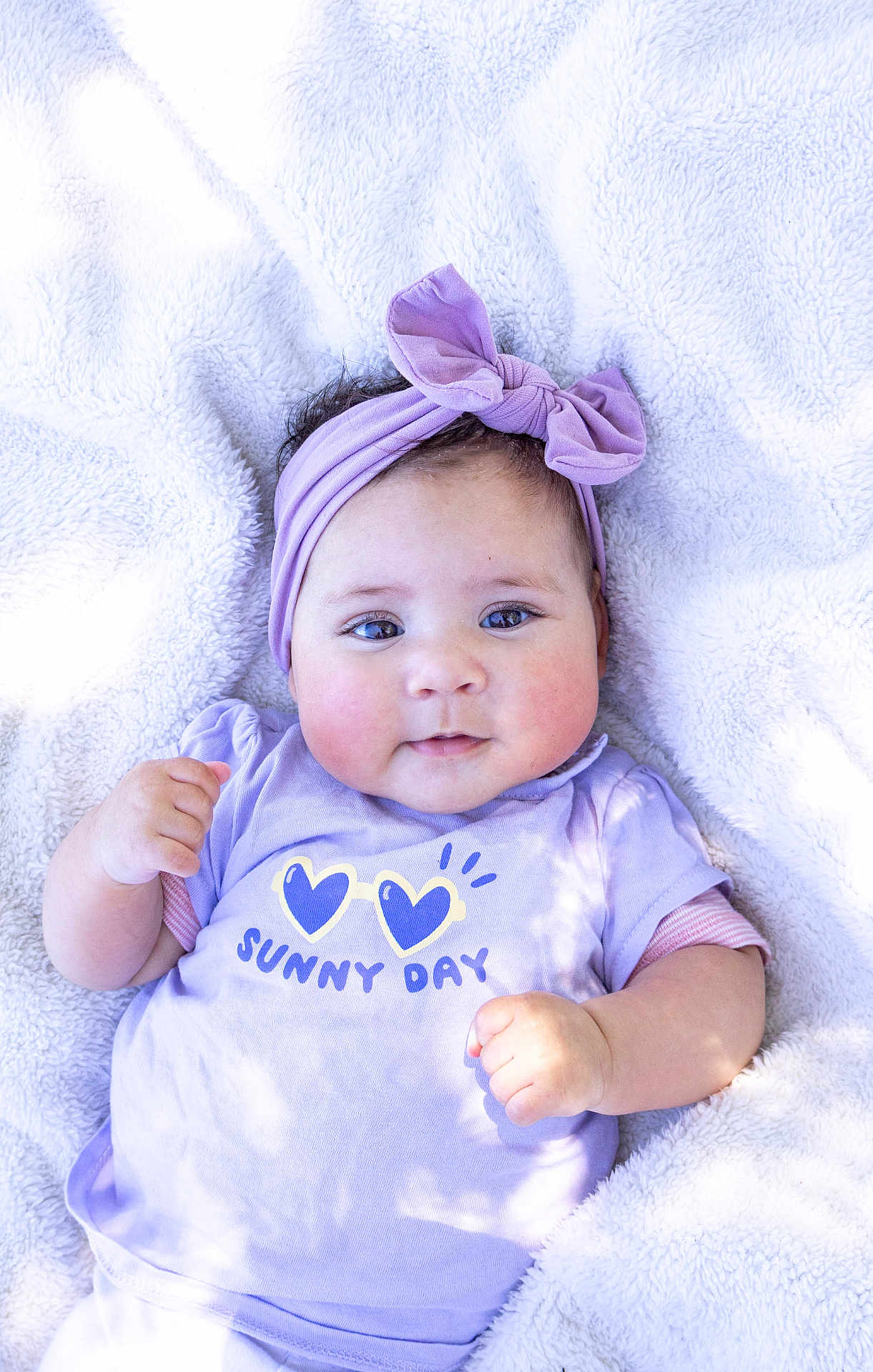 Manéa participe au concours pour gagner de l'argent avec cette photo : baby, blanket, bow, cheeks, child, cute, face, hand, headband, infant, lying_down, person, portrait, purple, shirt, skin, smile, soft_texture, sunny_day, text