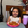 child, smiling, curly_hair, white_bow, plate, food, pink_cup, dining_table, chair, flowers, vase, vacuum_cleaner, home_interior, wooden_table, happy, girl, indoor, person, meal, cute