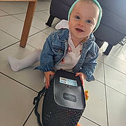 Zola a rejoint le concours — aidez-le/la à gagner de superbes lots ! toddler, child, indoor, floor, tile_floor, denim_jacket, headband, toy, basket, smiling, happy, person, sitting, furniture, couch, table_leg, white_tights, playful, cute, baby