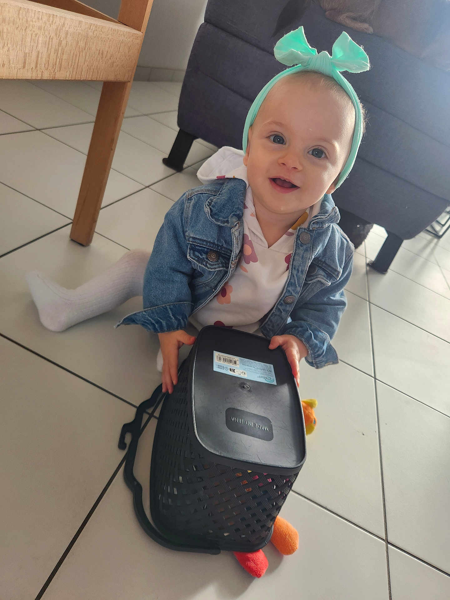 Zola a rejoint le concours — aidez-le/la à gagner de superbes lots ! toddler, child, indoor, floor, tile_floor, denim_jacket, headband, toy, basket, smiling, happy, person, sitting, furniture, couch, table_leg, white_tights, playful, cute, baby