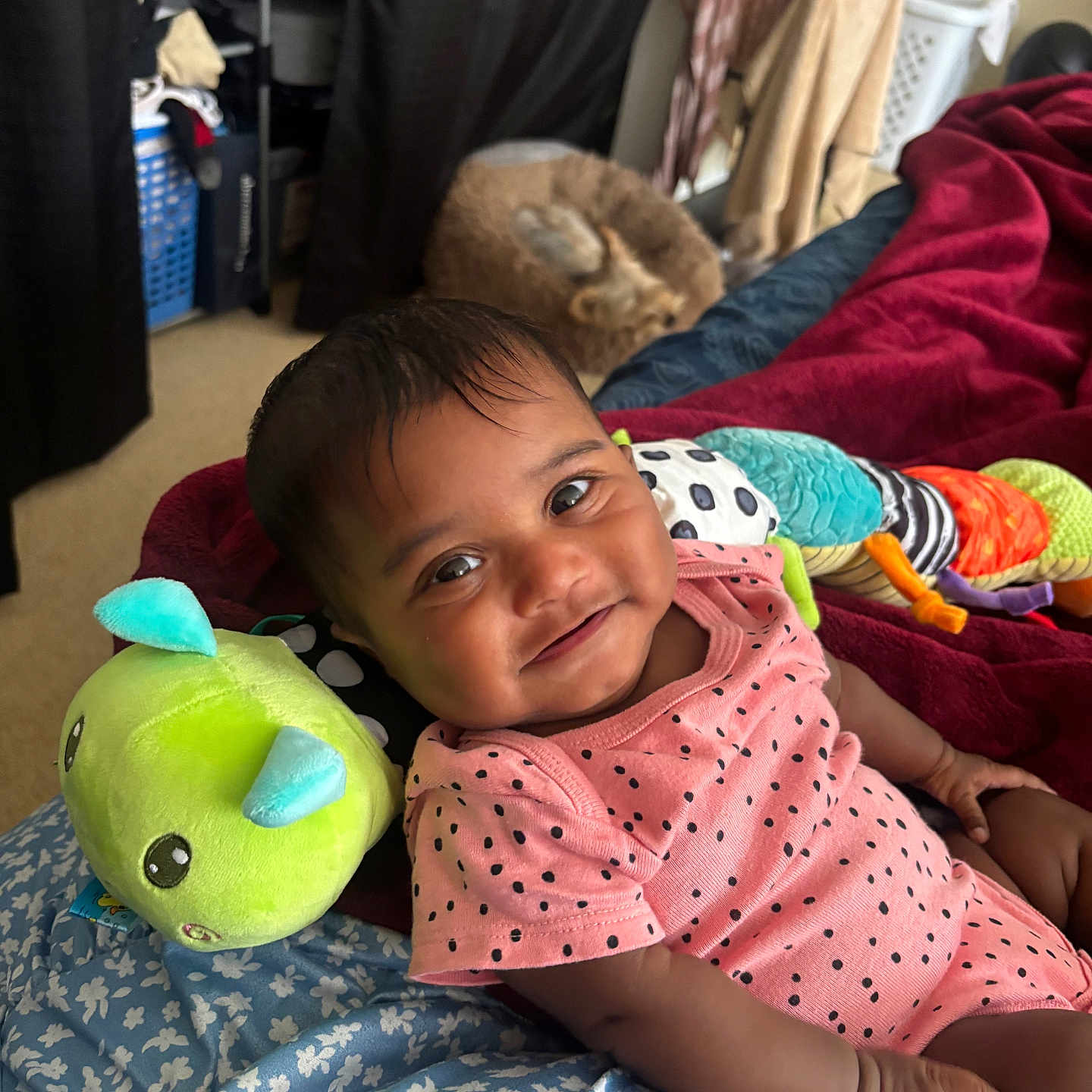 Ayra is registered to the contest to win money with this photo: baby, infant, smiling, pink_onesie, plush_toy, bed, blanket, room, laundry_basket, pet_bed, carpet, clothing, person, child, indoor, cute, happy, soft_toy, relaxing, home