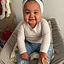 baby, child, smiling, headband, white_clothing, jeans, flower_patch, red_sandals, indoor, cushion, chair, cute, happy, portrait, person, small_child, clothing, furniture, baby_footwear, headwear