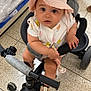baby, child, tricycle, hat, pink_hat, indoor, store, floor_tiles, footwear, onesie, handlebar, curious, seated, baby_clothing, toy_vehicle, pedals, shopping, baby_shoes, infant, cute