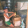 child, boy, duckling, cardboard_box, glasses, cap, green_shorts, white_tank_top, garage, storage, container, floor, bird, animal, smile, kneeling, indoor, clutter, toy_box, pet