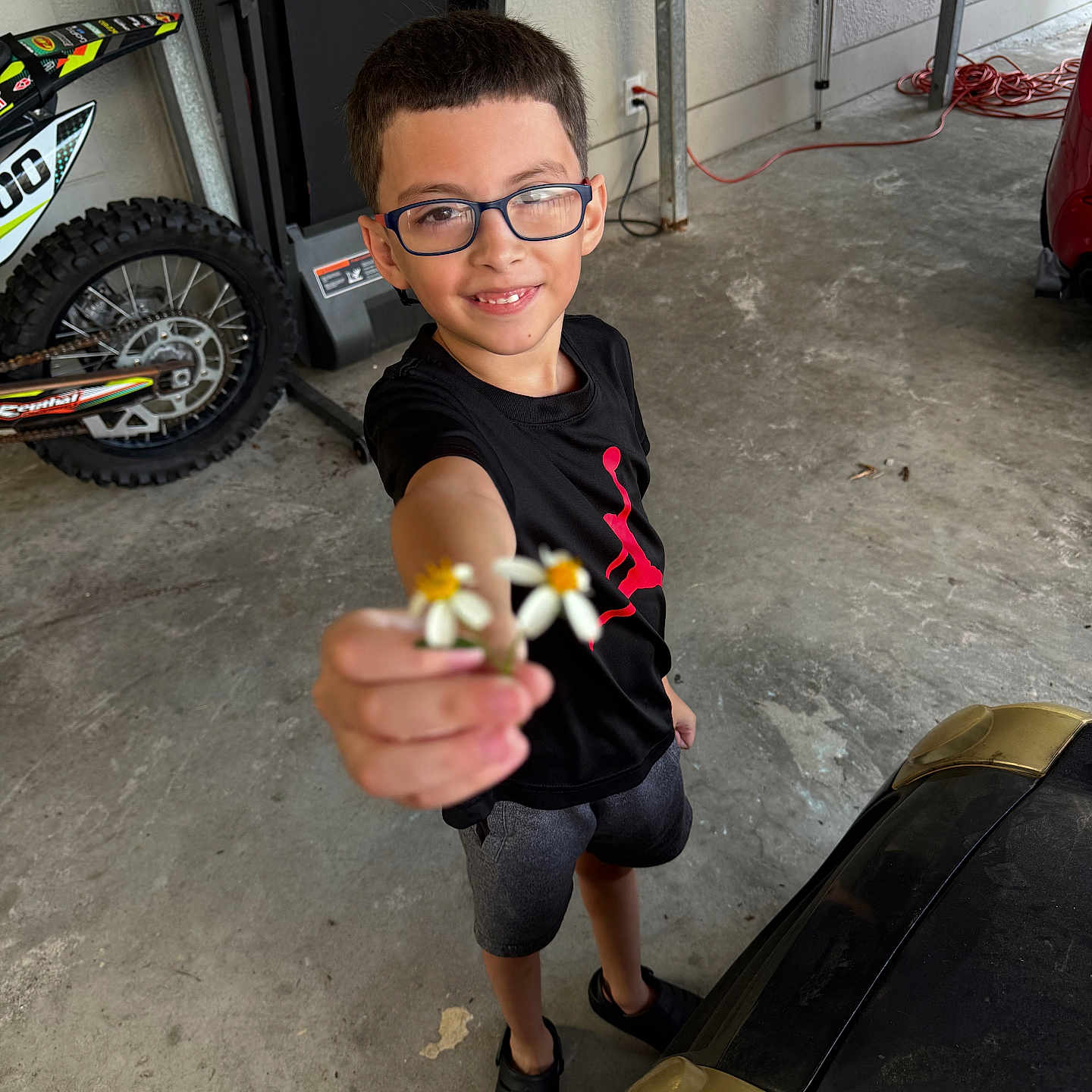 King is registered to the contest to win money with this photo: child, boy, glasses, smile, flowers, hand, black_shirt, shorts, garage, motorbike, concrete_floor, toy_car, cord, wall, footwear, person, young, casual_clothing, indoor, playful