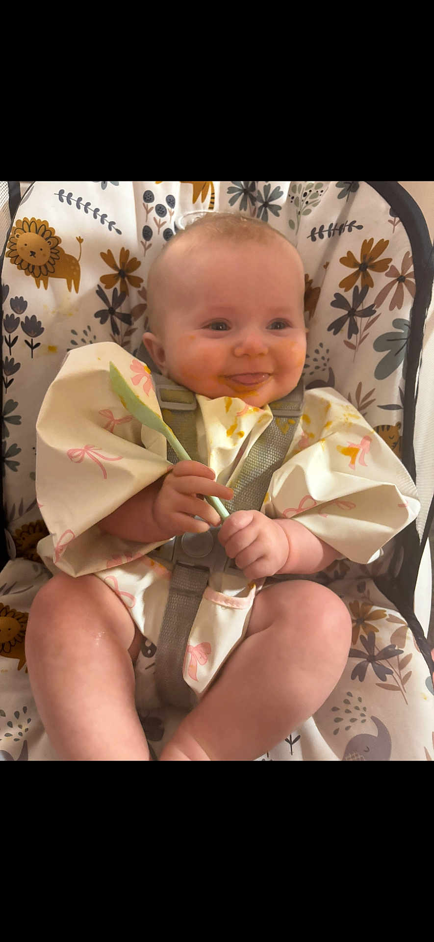 Bonnie is registered to the contest to win money with this photo: baby, child, high_chair, spoon, messy_face, smiling, food, feeding, infant, cute, playful_background, cartoon_animals, floral_pattern, clothing, sitting, happy, portrait, indoors, person, mealtime