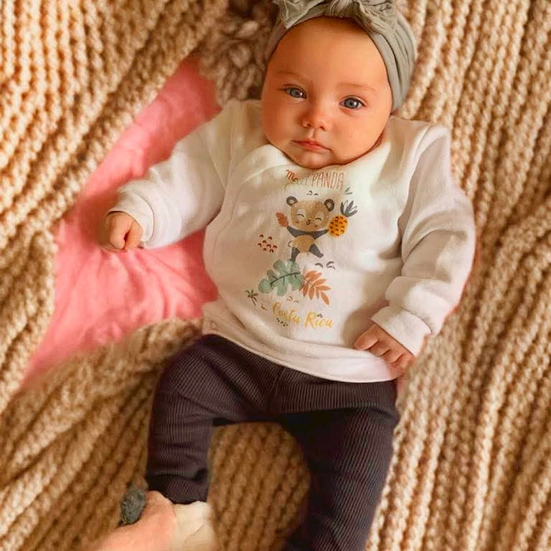 Jade a rejoint le concours — aidez-le/la à gagner de superbes lots ! baby, blanket, child, cozy, cute, dark_pants, face, feet, hands, headwrap, indoors, infant, knitted_blanket, lying_down, panda_print, pink_background, portrait, slippers, soft_lighting, white_shirt