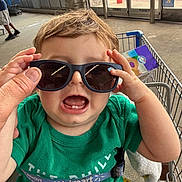 Vincent joined the competition — help win amazing prizes! child, toddler, baby, sunglasses, shopping_cart, supermarket, indoor, portrait, green_shirt, blond_hair, smiling, open_mouth, sitting, grocery_store, adult_hand, clothing, store_entrance, shopping, playful, photograph