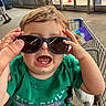 child, toddler, baby, sunglasses, shopping_cart, supermarket, indoor, portrait, green_shirt, blond_hair, smiling, open_mouth, sitting, grocery_store, adult_hand, clothing, store_entrance, shopping, playful, photograph