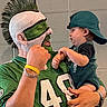 Vincent joined the competition — help win amazing prizes! baby, child, adult, father, portrait, football, football_jersey, sports_fan, face_paint, green_face, mohawk, backwards_cap, holding_child, indoor, brick_wall, family, smiling, costume, game_day, photograph