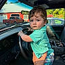 child, toddler, baby, boy, portrait, car, steering_wheel, driver_seat, adult_hand, outdoor, red_barn, pickup_truck, greenery, sunlight, blue_eyes, brown_hair, turquoise_shirt, plaid_shorts, vehicle_interior, family