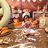 baby, child, baking, bread, cookie, flour, table, balloon, apron, chef_hat, wooden_spoon, rolling_pin, bakery, celebration, food, kitchen, smiling, cute, indoor, portrait
