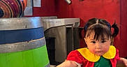 Annaya joined the competition — help win amazing prizes! barrel, black_shoes, checkered_floor, child, colorful_clothing, curious_expression, door, green_clothing, hand_reaching, high_chair, indoor, number_23, person, pigtails, red_wall, room, staircase, toddler, wooden_furniture, yellow_collar