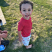 Xander is registered to the contest to win money with this photo: child, toddler, smiling, curly_hair, barefoot, red_shirt, white_shorts, lobster_print, grass, outdoor, sunlight, shadow, sidewalk, person, happy, playful, summer, casual_clothing, nature, daylight