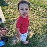 child, toddler, smiling, curly_hair, barefoot, red_shirt, white_shorts, lobster_print, grass, outdoor, sunlight, shadow, sidewalk, person, happy, playful, summer, casual_clothing, nature, daylight