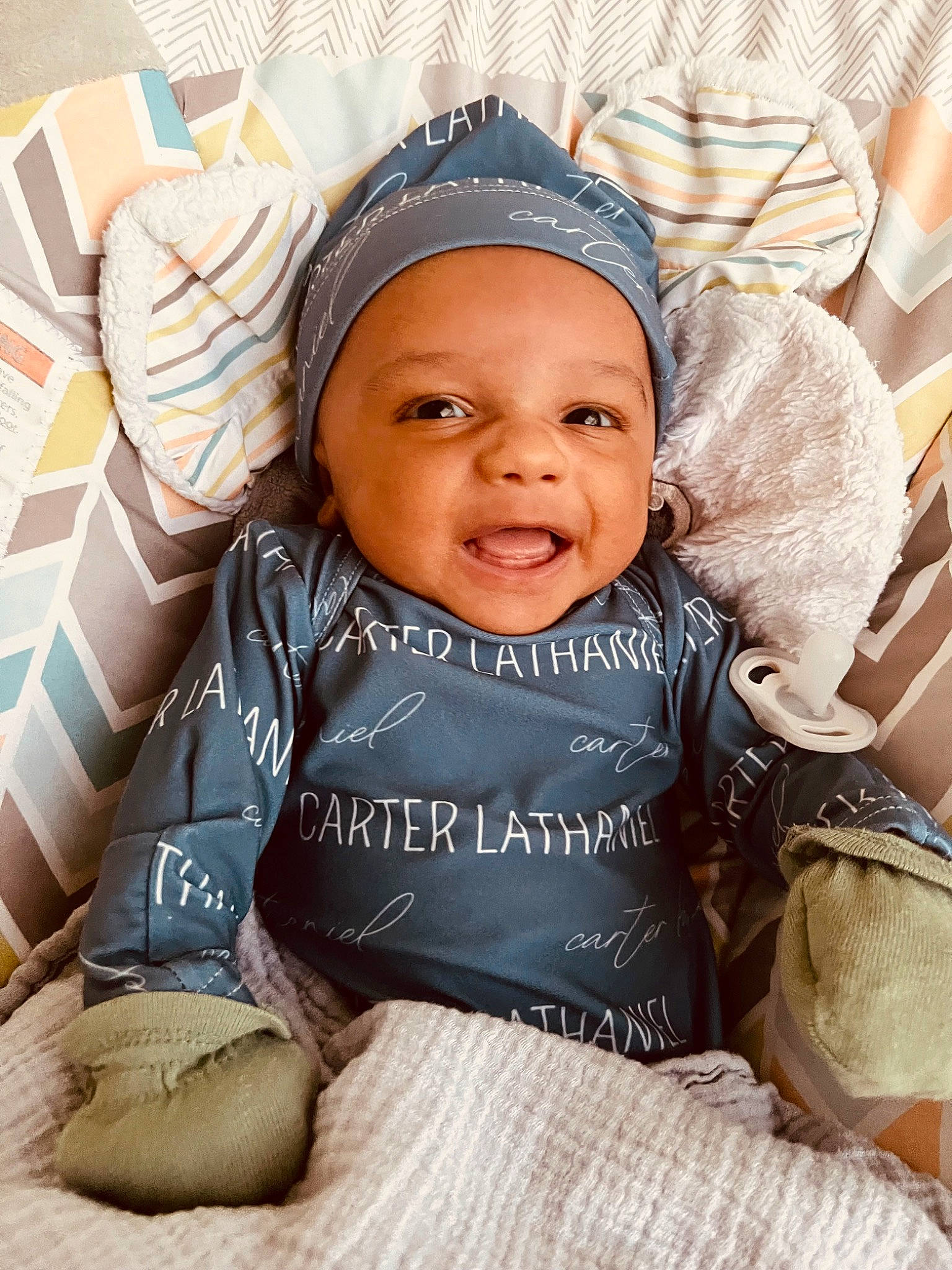 Carter is registered to the contest to win money with this photo: baby, baby_toddler_clothing, beanie, cap, chair, child, comfort, fashion_accessory, flooring, happy, headwear, linens, pattern, person, photo_caption, portrait_photography, room, sitting, sleeve, smile
