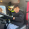 child, boy, smiling, car_interior, steering_wheel, seat, jacket, helmet, firefighter_gear, adult, man, seatbelt, water_bottle, vehicle, window, curly_hair, candid, portrait, happy, passenger
