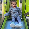 casual_clothing, child, face, green_play_structure, happy, jacket, metal_slide, outdoor, park, play_equipment, playground, portrait, sitting, slide, smiling, sneakers, socks, striped_leggings, toddler, young_child