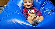 Troy is registered to the contest to win money with this photo: baby, child, bean_bag, blue, smiling, happy, indoor, clothing, striped_shirt, cute, person, face, socks, seated, furniture, wooden_door, floor, playful, cozy, comfort