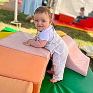 Gabin a rejoint le concours — aidez-le/la à gagner de superbes lots ! baby, child, outdoor, soft_play, play_blocks, colorful, smiling, curious, grass, padded_mats, person, clothing, white_shirt, overalls, barefoot, young_child, playground, daylight, people, fun