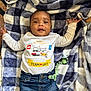 baby, infant, blanket, checkered, clothing, jeans, pacifier, face, person, lying_down, cute, child, young, indoor, smiling, arms_outstretched, blue, white, colorful_text, comfort