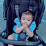 baby, stroller, blue_outfit, infant, hands_in_mouth, black_stroller, outdoor, pavement, cute, child, sitting, curious_eyes, baby_boy, baby_girl, daylight, casual, child_in_stroller, baby_feet, baby_face, childhood