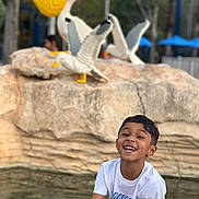 Rhylan Sarjoo joined the competition — help win amazing prizes! child, boy, smile, laughing, fountain, water, rock, sculpture, pelican, birds, lego_style, outdoor, park, trees, blurred_background, portrait, white_shirt, nike_logo, happy, playful