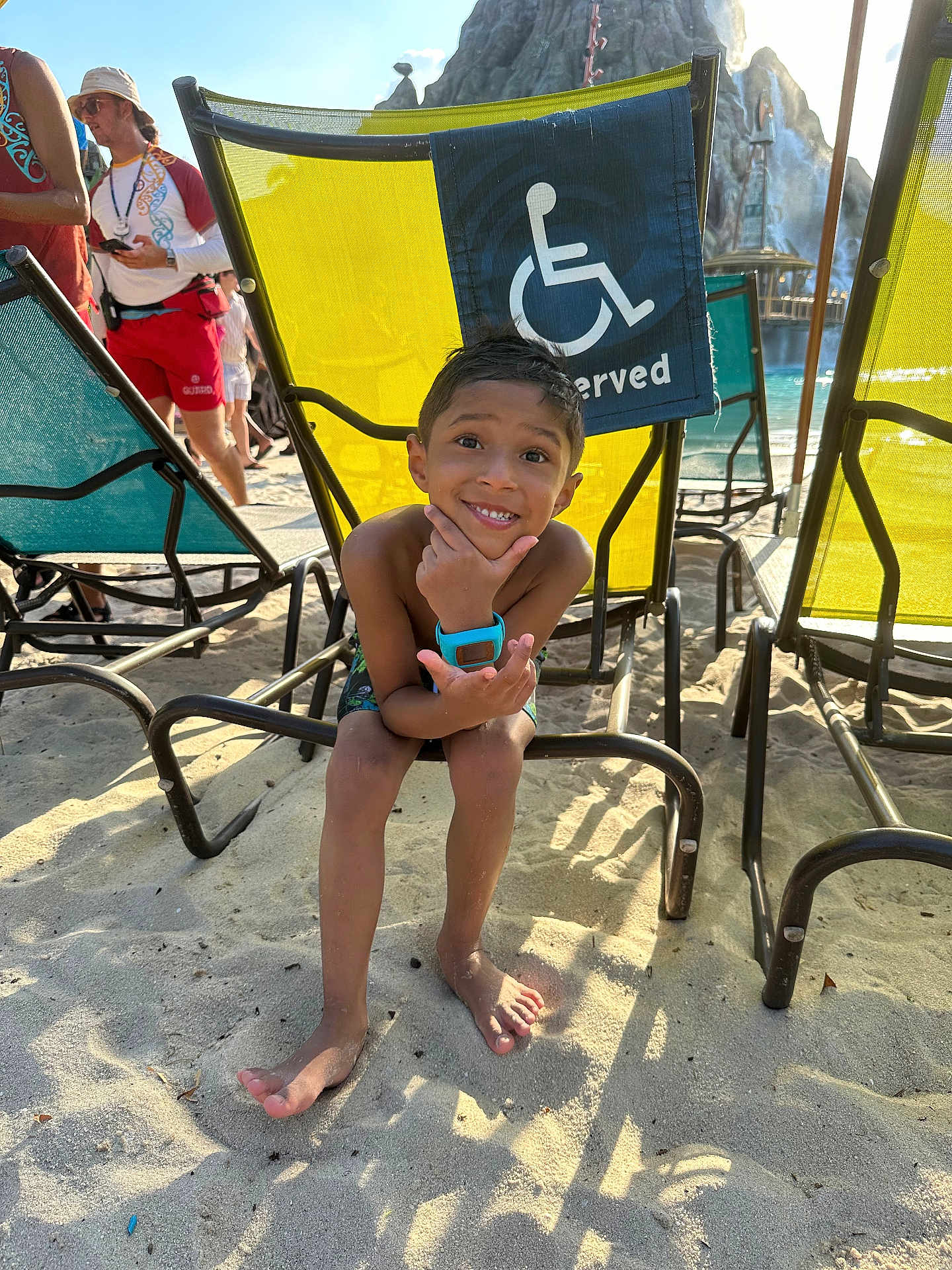 Rhylan Sarjoo is registered to the contest to win money with this photo: child, boy, smiling, beach, sand, lounge_chair, reserved_sign, wheelchair_symbol, swim_shorts, turquoise_watch, barefoot, lifeguard, sunglasses, waterpark, pool, vacation, summer, playful_pose, outdoor, family