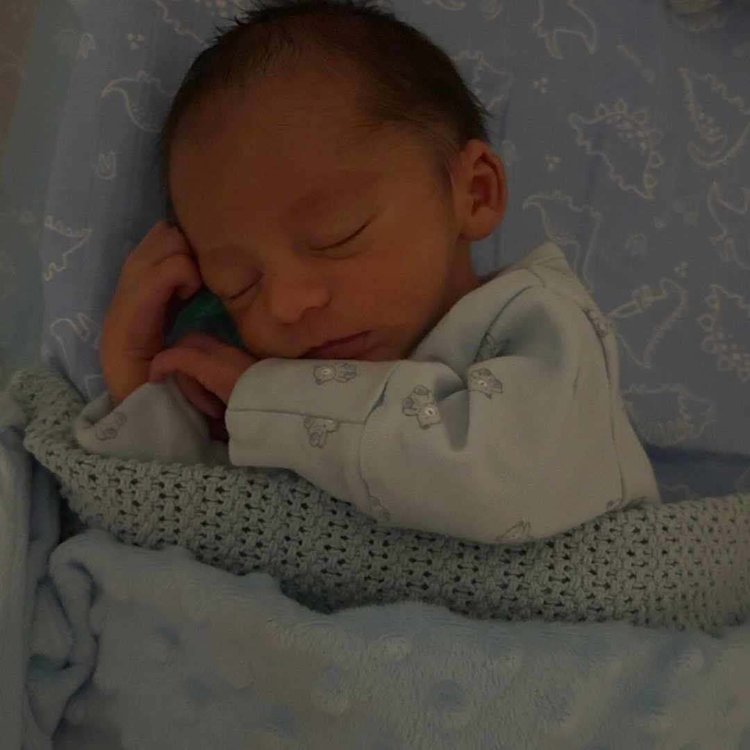 Noah is registered to the contest to win money with this photo: baby, bed, blanket, blue, child, cozy, cute, dinosaur_pattern, head, indoors, infant, newborn, pajamas, peaceful, person, portrait, resting, sleeping, soft, toddler