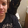 person, cat, black_cat, indoor, selfie, stool, yellow_eyes, concerned_expression, textured_wall, casual_clothing, black_shirt, chain_necklace, pet, animal, portrait, close_up, feline, home, fur, whiskers