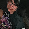 person, glasses, smile, selfie, black_cat, cat, pet, closeup, indoors, bed, blanket, tshirt, graphic_tee, face, eyewear, feline, cozy, resting, dark, hair