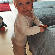 Lyam participe au concours pour gagner de l'argent avec cette photo : toddler, child, indoor, blanket, furniture, toy, penguin_toy, floor, curtains, red_rug, white_shirt, pants, socks, standing, looking_at_camera, baby, light, window, home, portrait
