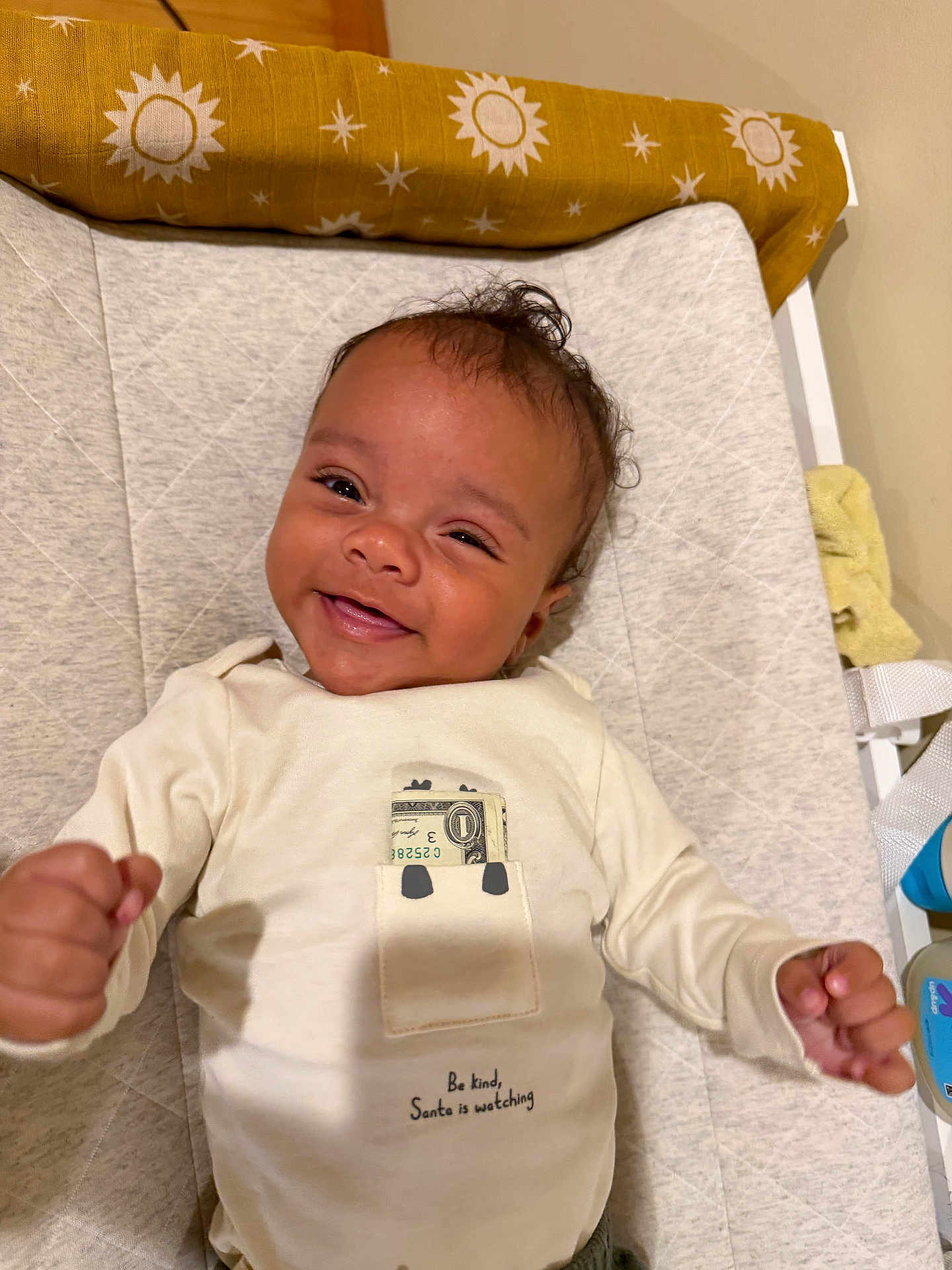 Marley is registered to the contest to win money with this photo: baby, smiling, infant, onesie, dollar_bill, pocket, text_on_clothing, changing_pad, raised_fists, happy, indoor, fabric, pattern, baby_care, cute, toddler, child, face, person, clothing