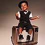 black_clothing, bow_tie, child, cute, fashion, happy, hat, minimalist_background, playful, portrait, retro, shoes, shorts, sitting, smiling, socks, studio_lighting, toddler, vintage_television, wooden_tv