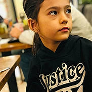 Arya is registered to the contest to win money with this photo: child, girl, portrait, hoodie, black_clothing, indoor, table, blurred_background, face, looking_away, casual, young, person, expression, earrings, brown_hair, sitting, restaurant, wooden_table, soft_lighting