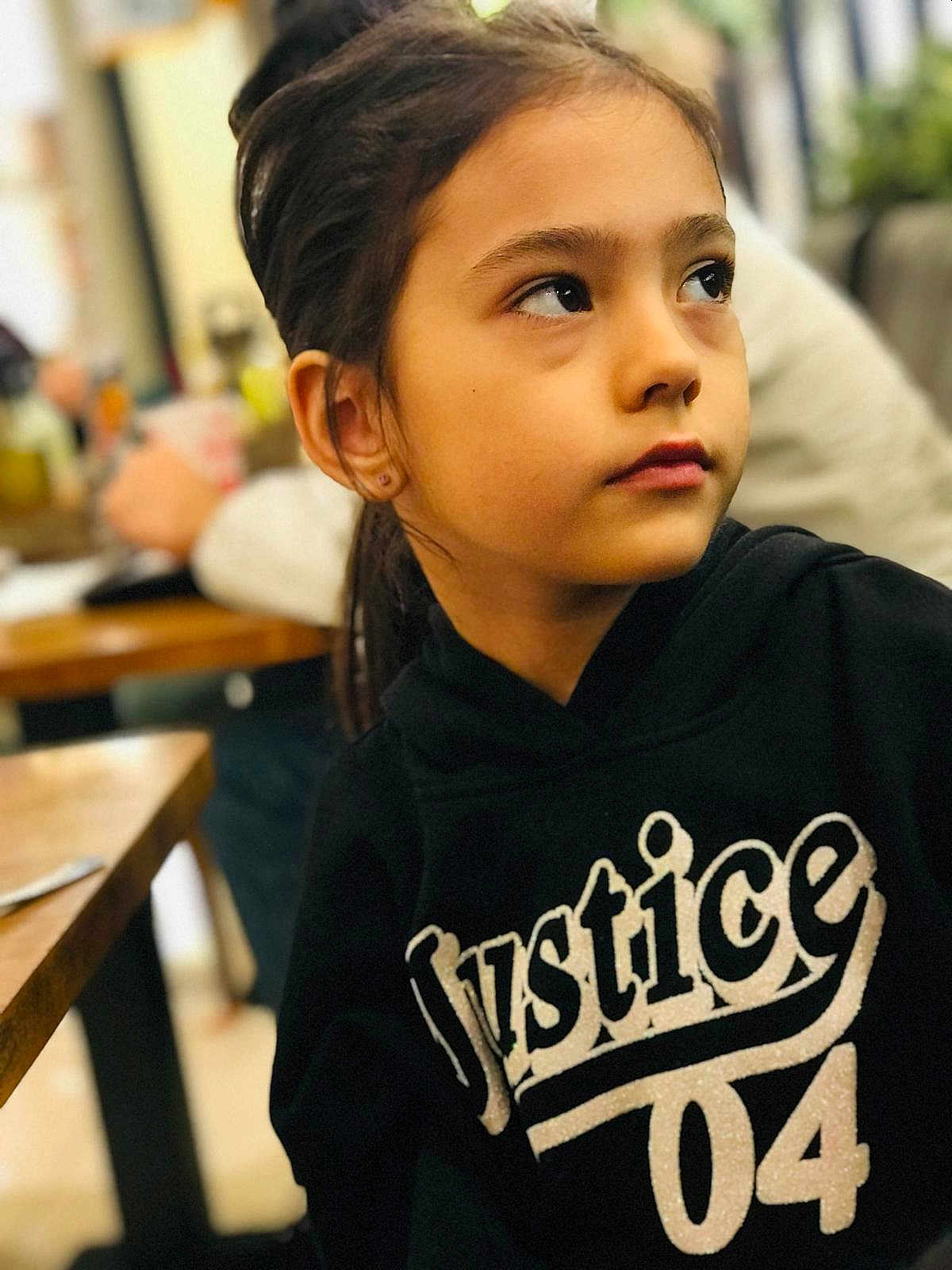 Arya is registered to the contest to win money with this photo: child, girl, portrait, hoodie, black_clothing, indoor, table, blurred_background, face, looking_away, casual, young, person, expression, earrings, brown_hair, sitting, restaurant, wooden_table, soft_lighting