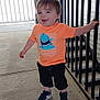 toddler, child, smiling, orange_tshirt, shark_graphic, shorts, sneakers, black_shoes, standing, railing, porch, outdoor, concrete_floor, happy, casual_clothing, person, young_child, short_hair, daylight, holding