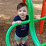 toddler, child, playground, play_equipment, green_bars, shorts, sneakers, wood_chips, outdoor, daylight, curious, standing, casual_clothing, short_hair, smiling, young_child, park, fun, play, person