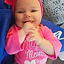 baby, baby_girl, blue_fabric, child, clothing, cushion, cute, face, hand_in_mouth, happy, headwear, indoors, infant, patterned_background, person, pink_headband, pink_shirt, portrait, sitting, smiling